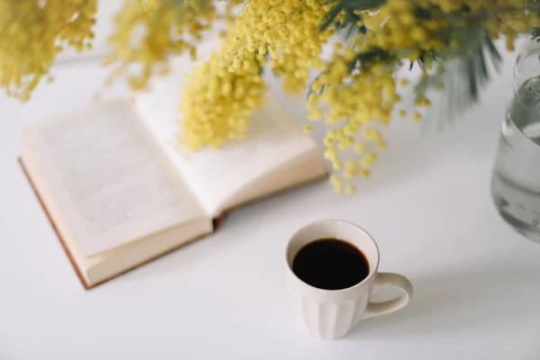 Xícara de café e um livro sobre mesa branca com flores amarelas bouquet mimosa. Conceito de planejamento e design. Local de trabalho. Instagram deitado plano feminino. Vista superior. Pequeno-almoço acolhedor. Páscoa, conceito de primavera — Fotografia de Stock