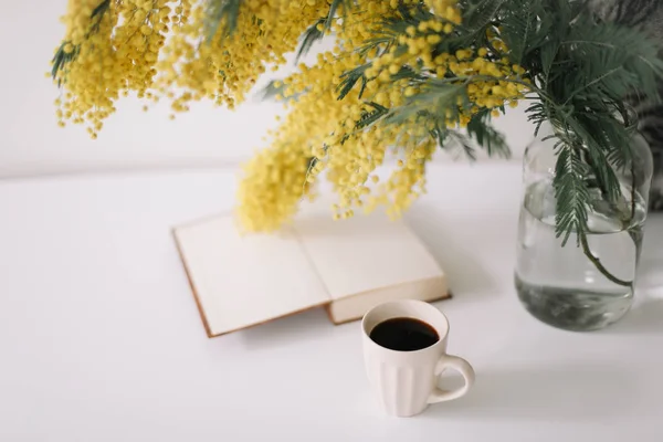 Xícara de café e um livro sobre mesa branca com flores amarelas bouquet mimosa. Conceito de planejamento e design. Local de trabalho. Instagram deitado plano feminino. Vista superior. Pequeno-almoço acolhedor. Páscoa, conceito de primavera — Fotografia de Stock
