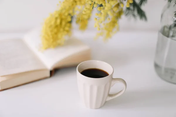 Kopp kaffe och en bok på vitt skrivbord med gula blommor bukett mimosa. Planering och design koncept. Arbetsplatsen. Instagram feminina platt lay. Ovanifrån. Mysig frukost. Påsk, våren koncept — Stockfoto