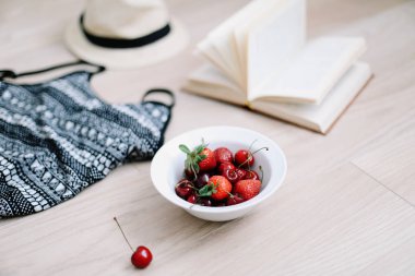 En iyi görünüm seyahat veya tatil konsepti. Mayo, şapka, kitap ve ahşap arka plan üzerinde taze tatlı kiraz ve çilek. Düzlay. Yaz arka planı. 