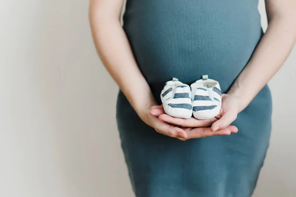 Mother hands holding shoes for the newborn baby. Pregnant woman touching belly. Happy woman pregnancy, maternity, body care