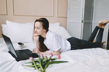 Yatakta beyaz çarşaflarla dizüstü bilgisayarda çalışan genç bir kadın. Yaşam tarzı kompozisyonu. Evde çalışmak, karantina kavramı. 