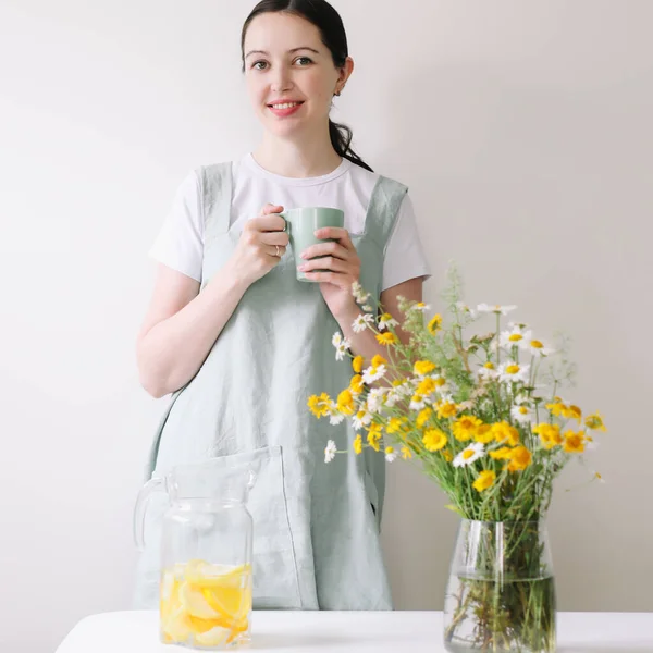 Nő vászon kötényben limonádét iszik kényelmes otthoni háttérrel. Tiszta természetes szövet kötény. Élelmiszer blogger, házi készítésű főzés — Stock Fotó