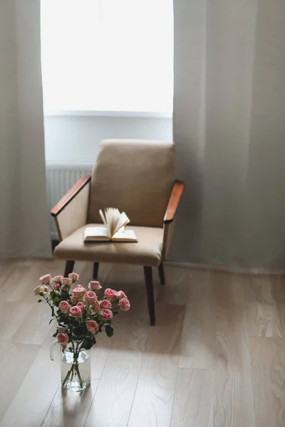 Moderne kamer interieur met fauteuil, boek en verse rozen op tafel. Interieur van mooie woonkamer versierd met bloemen — Stockfoto