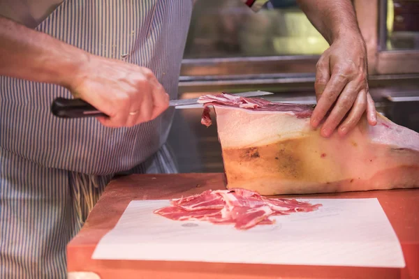 ハモン ブッチャー ナイフで肉を切る 肉屋が見られます ハモンは新鮮に見えるし カットされた後販売すること準備ができています ストライプのエプロンを着て見られています — ストック写真