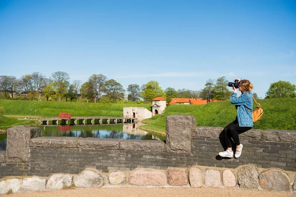 Young woman with camera sitting on stone fence and photographing Citadel, copenhagen, denmark — Stock Photo