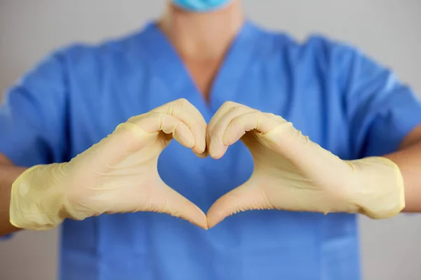 Doctor or nurse in a blue shirt makes the heart symbol with hands in gloves.
