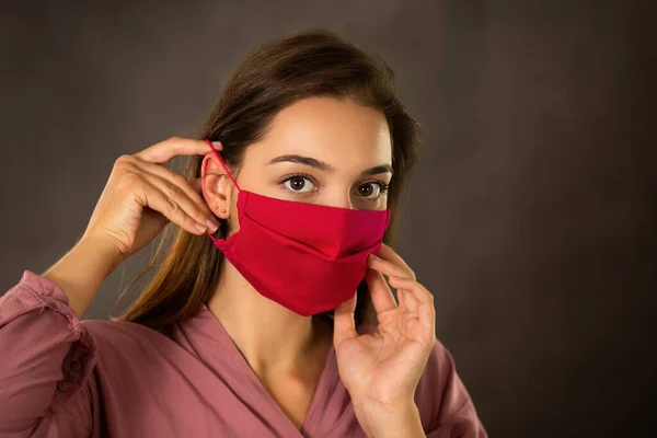 Woman attaching red cloth face mask on ear with fingers with dark background — Stock Photo, Image