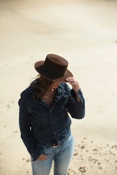 穿着棕色帽子和牛仔裤夹克的黑发女人站在沙漠里 — 图库照片