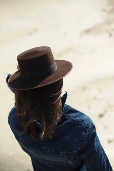 穿着棕色帽子和牛仔裤夹克的黑发女人站在有风的沙漠里 后视图 — 图库照片