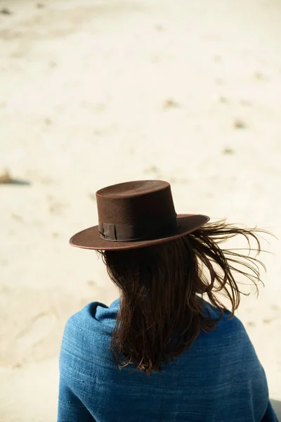 棕色帽子和蓝色布的黑发妇女站立在有风的沙漠 后视图 — 图库照片