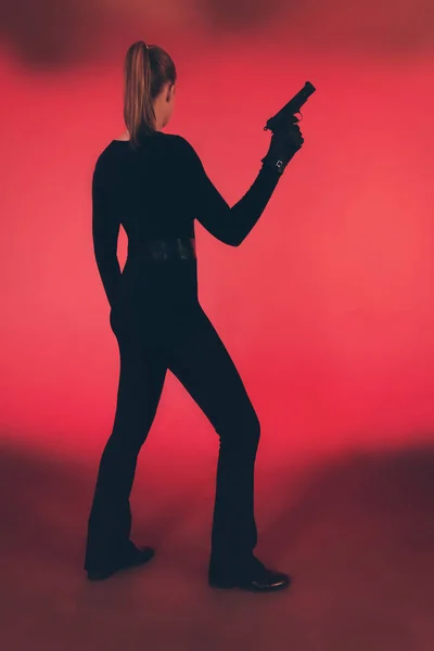 Silhouette of woman in black holding pistol. Against red backgro