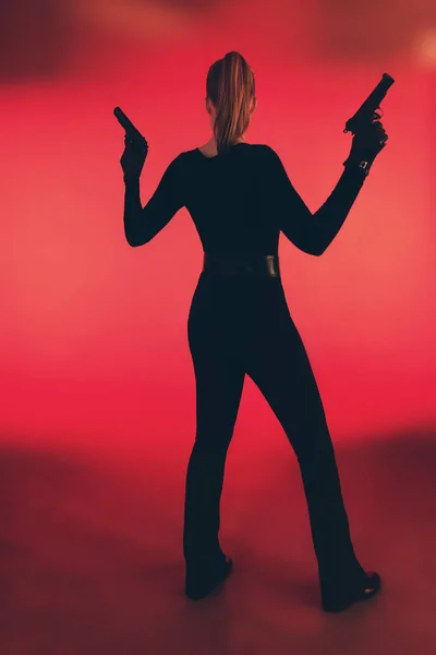 Menina com duas armas vestidas de preto. De pé contra o backgr vermelho — Fotografia de Stock