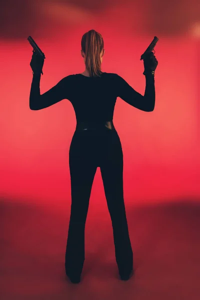 Girl with two guns dressed in black. Standing against red backgr — Stock Photo, Image