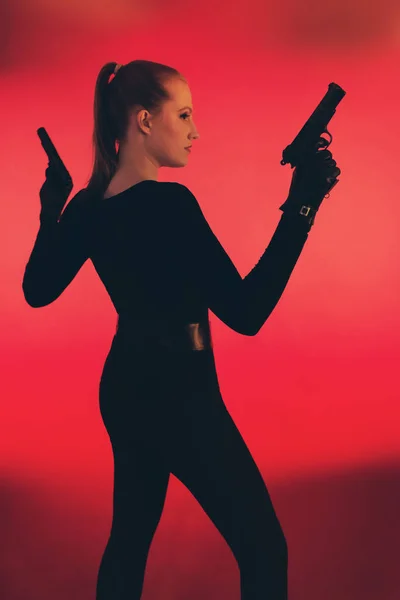 Girl with two guns dressed in black. Standing against red backgr — Stock Photo, Image