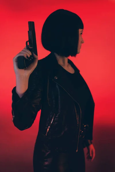 Woman with gun dressed in black against red background.
