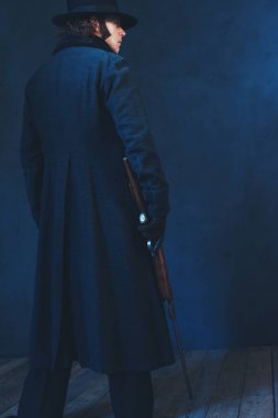 Uzun siyah ceket ve şapka tutan tüfekli Victorian adam. Arka Vie