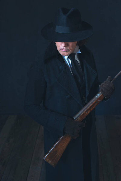 Victorian man in long black coat and hat holding rifle.