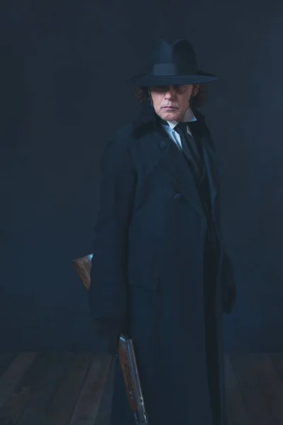 Victorian man in long black coat and hat holding rifle.