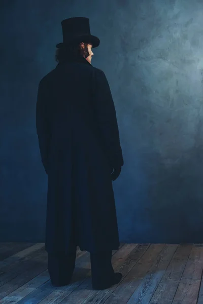 Uomo edoardiano in lungo cappotto nero e cappello in piedi verso il grigio w — Foto Stock