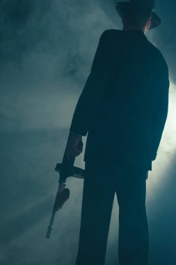 Retro gangster dumanlı odada makineli tüfek ile duruyor. Arka görüş.