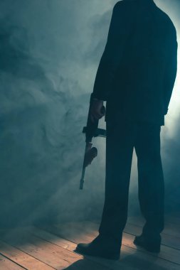 Retro gangster dumanlı odada makineli tüfek ile duruyor. Arka görüş.