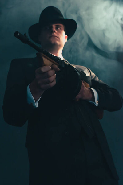 Retro gangster in hat in misty night shoots with machine gun.