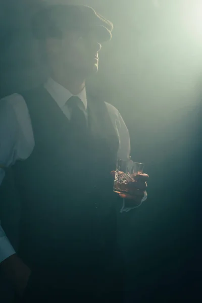 Homme d'affaires rétro avec capuchon tient un verre de whisky dans une pièce fumée . — Photo