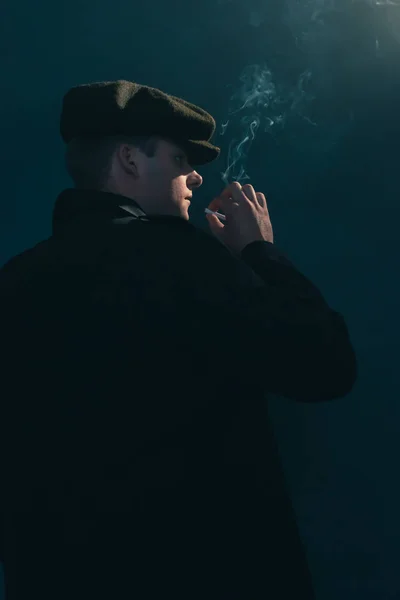Retro man with cap smokes cigarette in smoky room. Side view.