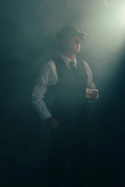 Homme d'affaires rétro avec capuchon tient un verre de whisky dans une pièce fumée . — Photo