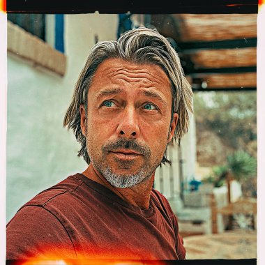 Bronz tenli, orta yaşlı, sarışın ve sakallı bir adam bahçede beyaz İspanyol evinin yanında oturuyor..