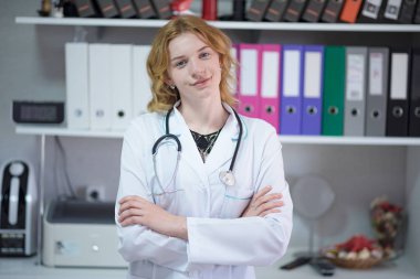 Tell me about your illnesses. Portrait of beautiful blonde female doctor in office with laptop. She sits right in front of the camera smiling and looks serious clipart