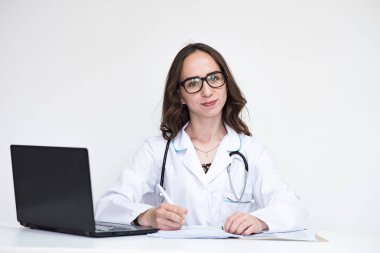 Hastalıklar hakkında söyle. Dizüstü bilgisayar ile beyaz arka plan üzerinde güzel esmer doktor kız portresi. O şu kamera gülümseyen önünde oturur ve ciddi görünüyor