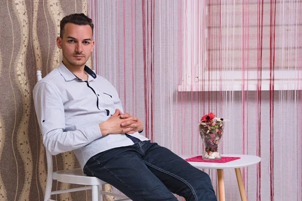 Portrait of a cute young man sitting on a chair in a room. He sits right in front of the camera and looks serious.