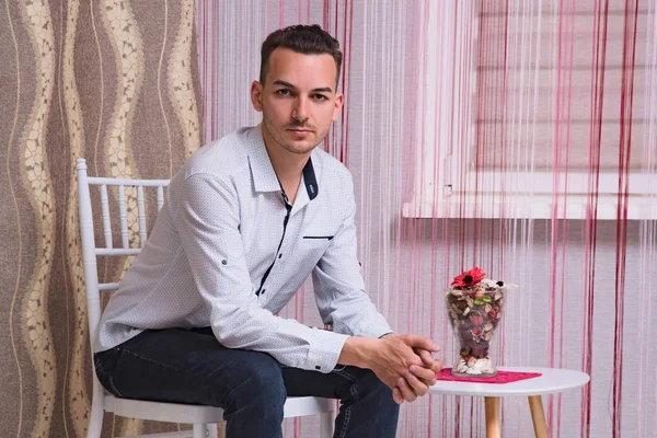 Portrait of a cute young man sitting on a chair in a room. He sits right in front of the camera and looks serious.