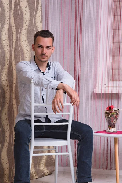 Portrait of a cute young man sitting on a chair in a room. He sits right in front of the camera and looks serious.