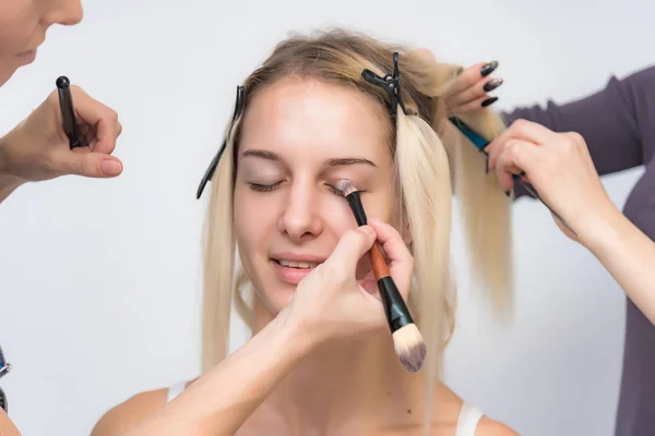 The work of a professional makeup artist - beautician, makes makeup with a brush on the face of a beautiful blonde with shadows on the eyes of the model. Painting of eyebrows.