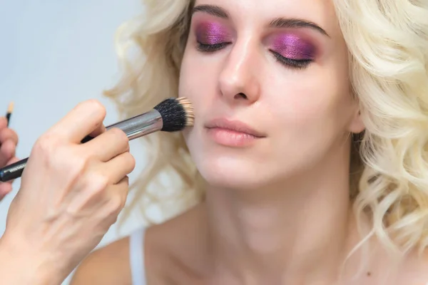 The work of a professional makeup artist - beautician, makes makeup with a brush on the face of a beautiful blonde with shadows on the eyes of the model. Painting of eyebrows.