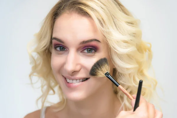 The work of a professional makeup artist - beautician, makes makeup with a brush on the face of a beautiful blonde with shadows on the eyes of the model. Painting of eyebrows.