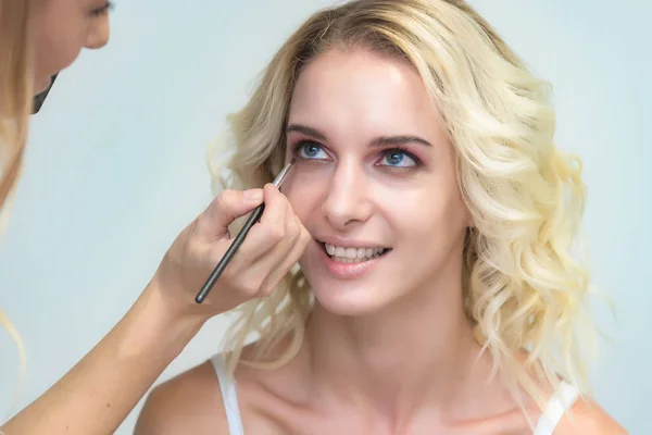 The work of a professional makeup artist - beautician, makes makeup with a brush on the face of a beautiful blonde with shadows on the eyes of the model. Painting of eyebrows.