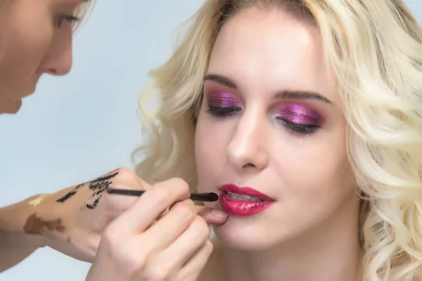 The work of a professional makeup artist - beautician, makes makeup with a brush on the face of a beautiful blonde on the lips of a model. Makeup artist creating beautiful makeup for blonde model