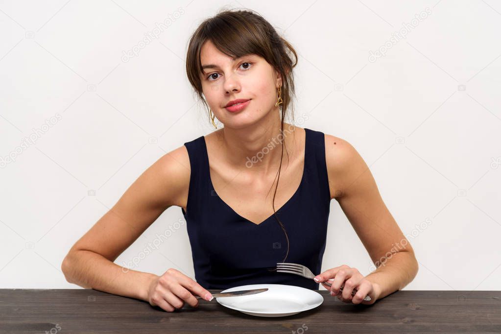Studio photo portrait of a beautiful brunette girl on a white background sitting at a table with a plate for food. She sits right in front of the camera in various poses, smiling and looking happy.