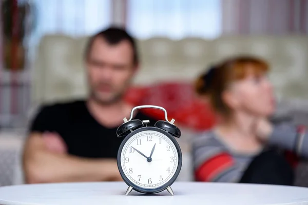 husband and wife are in the room and fighting,they are sitting on the bed, and angry, in the foreground of the clock, they are in the middle of the frame and look at the camera.