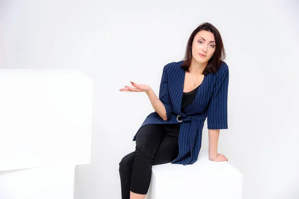 Concept portrait of a girl sitting on a white background in a business suit in different poses with different emotions. It in the middle of the frame, next to the white cubes, next is another cube.