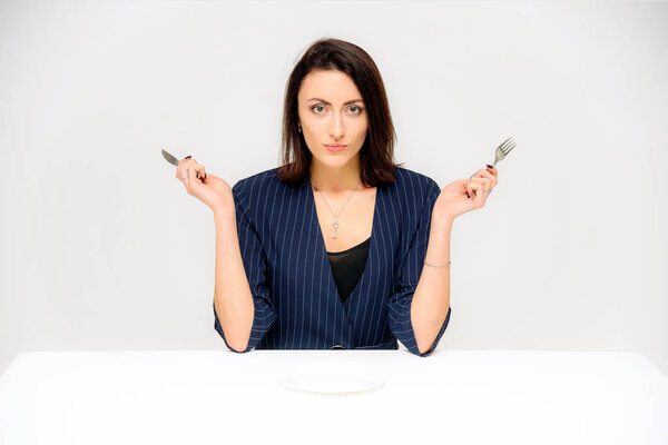 Концептуальный портрет брюнетки, сидящей на белом фоне в деловом костюме с тарелкой, вилкой, ложкой. Она сидит прямо перед камерой в разных позах с разными эмоциями
.