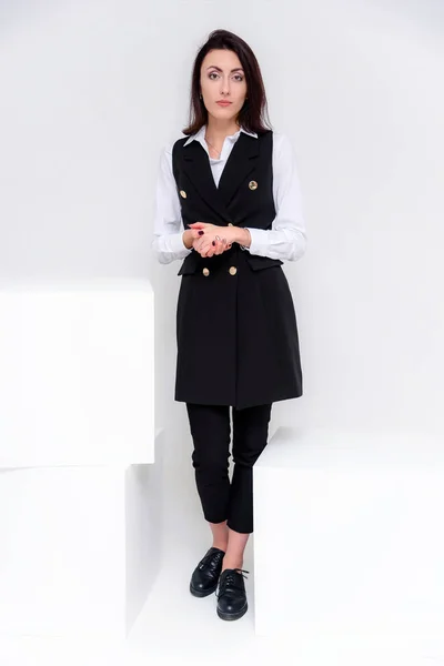Concept portrait of a girl standing on a white background in a business suit in different poses with different emotions. It in the middle of the frame, next to the white cubes, next is another cube.