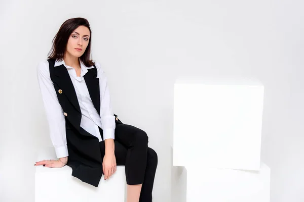 Concept portrait of a girl sitting on a white background in a business suit in different poses with different emotions. It in the middle of the frame, next to the white cubes, next is another cube.