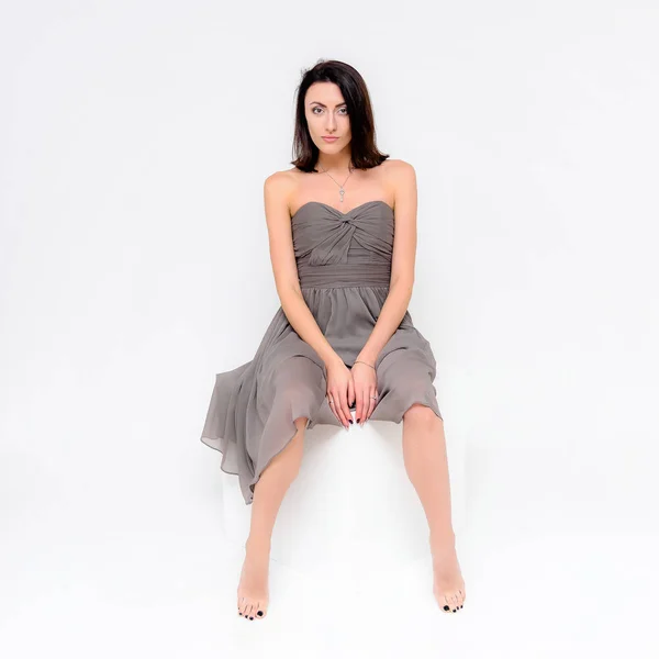 Concept portrait of a brunette girl, sitting on a white background in a beautiful brown dress on a white cube in studio. She sits right in front of the camera in various poses with different emotions.