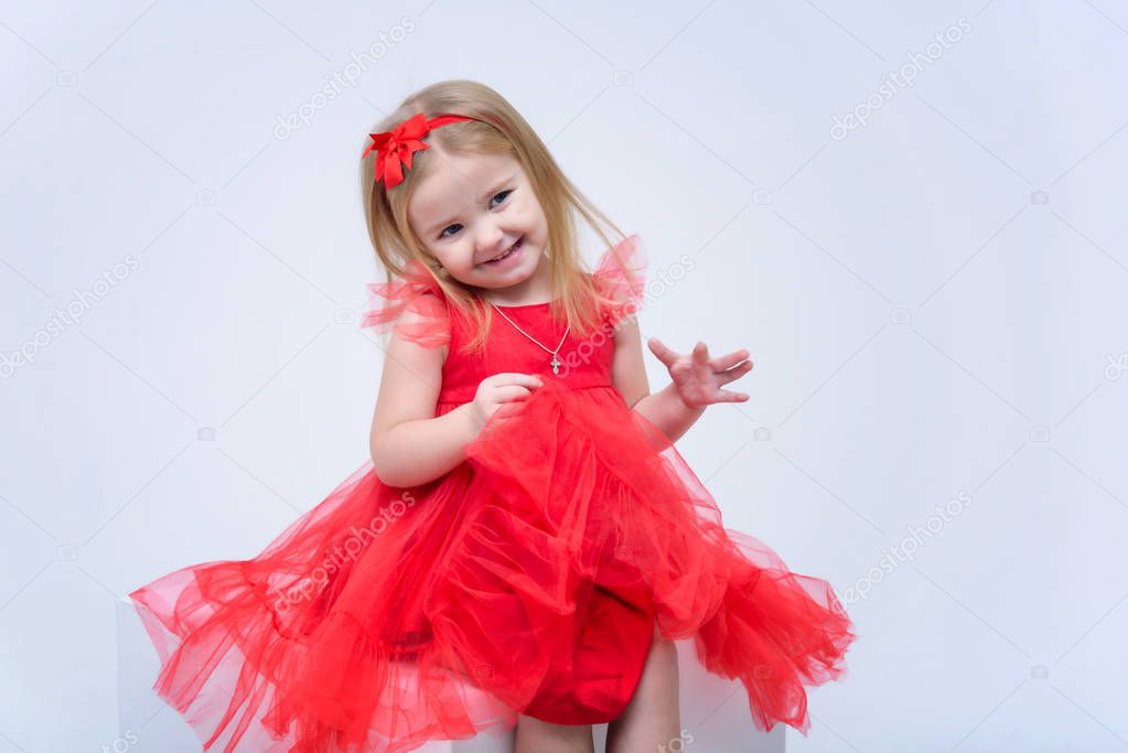 Concept portrait of a cute beautiful baby girl on a white background in a fashionable red dress. She sits on a cube right in front of the camera in various poses and shows different emotions.