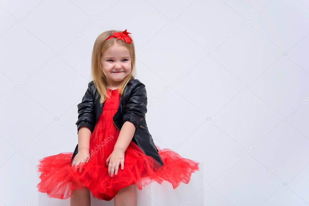 Concept portrait of a cute beautiful baby girl on a white background in a fashionable red dress. She sits on a cube right in front of the camera in various poses and shows different emotions.